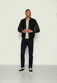 Man wearing black bomber jacket with leather sleeves, gray shirt, dark jeans, and black-and-white sneakers, standing against plain background.
