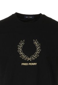 Camiseta negra de algodón con una corona de laurel bordada en dorado y el texto "FRED PERRY" debajo del diseño. Cuello redondo clásico.