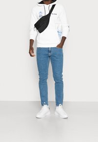 Sudadera blanca con gráficos azules, jeans azules y zapatillas blancas. Se lleva un bolso cruzado negro sobre el pecho.