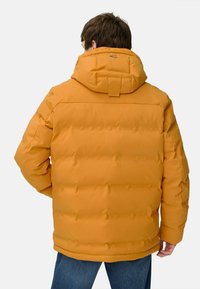 Mand iført en sennepsgul pufferjakke med hætte, vist fra ryggen, parret med blå jeans mod en ensfarvet baggrund.