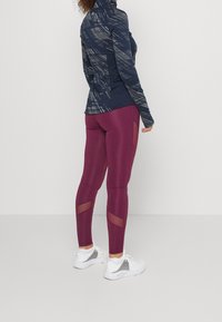 Sportig outfit bestående av en navy-mönstrad långärmad topp och vinröda leggings med mesh-paneler, matchat med vita sportskor.