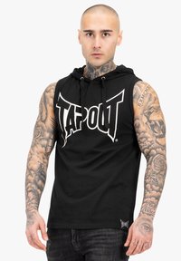 Melna bezpiedurkņu kapuce ar "TAPOUT" logo baltā krāsā, regulējams aukliņas aizdare un teksturēta auduma. Redzami ievērojami tetovējumi uz rokām.