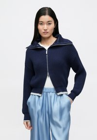 Suéter de punto azul marino con cremallera, textura acanalada y cuello plegable. Detalles en gris claro en los puños y el dobladillo. Combinado con pantalones azul claro.