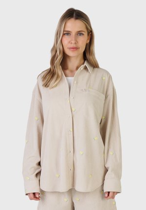 Frau mit blonden, welligen Haaren trägt ein beiges Hemd mit Knöpfen und passende Shorts mit kleinen gelben Zitronenmotiven auf hellem Hintergrund.