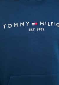 Modrá bavlněná mikina s kapucí s předním logem "TOMMY HILFIGER EST. 1985", která má červený, bílý a modrý obdélníkový akcent. Měkká textura.