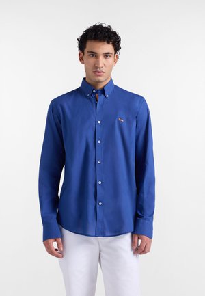 Blaues Hemd aus glattem Stoff mit langen Ärmeln, das ein kleines oranges Logo auf der linken Brust und weiße Knöpfe aufweist.