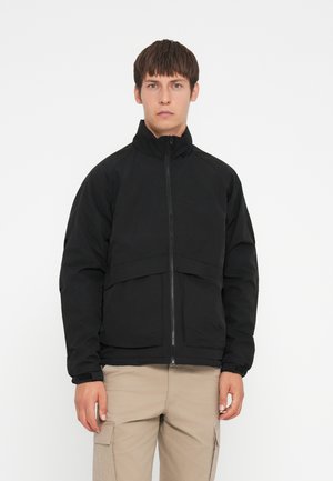 Black zip-up jacket with a high collar, featuring a smooth texture, structured design, and subtle seam details, worn over khaki pants.