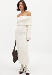 Abito lavorato a maglia off-the-shoulder in crema, silhouette aderente, scollo a coste, maniche lunghe, spacco laterale, abbinato a una borsa marrone texturizzata e calze beige.