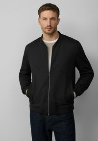 Veste bomber noire avec fermeture éclair sur le devant, col et poignets côtelés. Comprend deux poches latérales et une texture de tissu lisse. Portée par-dessus une chemise légère.