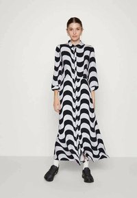 Vestido longo com padrão ondulado em preto e branco, com colarinho, botões à frente e mangas até ao cotovelo. A modelo está a usar sapatos pretos.