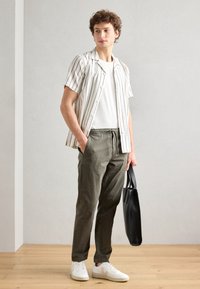 Cercel cu mânecă scurtă, cu dungi albe și gri, asortat cu pantaloni verde măsliniu și adidași albi, ținând o geantă de mână neagră.
