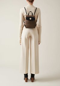 Brown textured backpack with black accents and a circular gold clasp, worn on a person’s back, with wide cream-colored trousers.