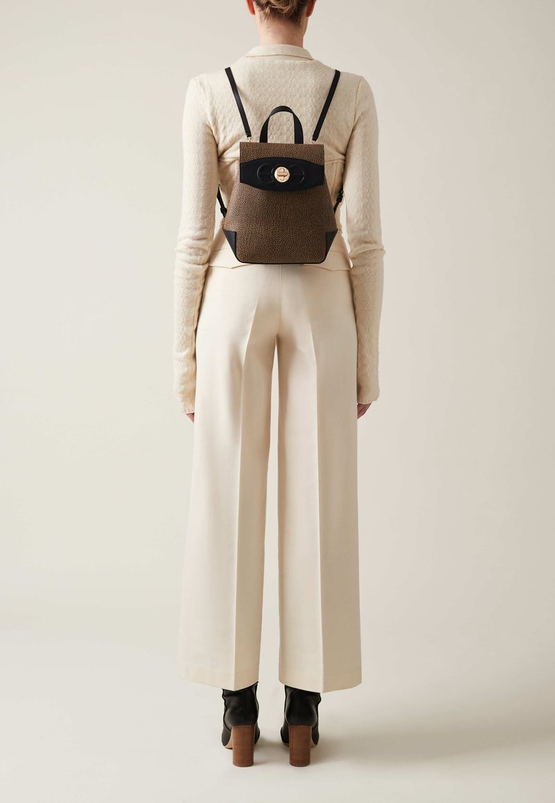 Brown textured backpack with black accents and a circular gold clasp, worn on a person’s back, with wide cream-colored trousers.