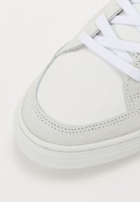 Hvit sneakers med overdel i semsket og glatt skinn, rund tå, hvite snører og teksturert gummisåle. Subtile sømdetaljer er synlige.