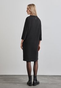 Schwarzes, knielanges Kleid mit Dreiviertelärmeln, strukturiertem Material und einem kleinen Logodetail auf der Rückseite. Kombiniert mit schwarzen Stiefeletten.