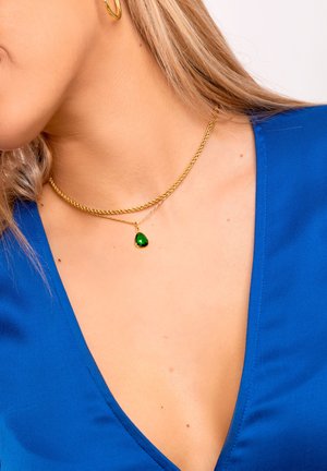 Collane a strati in tono oro con una catena attorcigliata e un pendente in forma di goccia di pietra verde, poggiato su una maglietta blu.