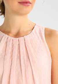 Detailní záběr na krk a rameno ženy, která nosí bezrukávový, světle růžový strukturovaný top s záhyby u límce.