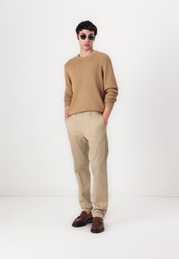 Beige žebrovaný svetr, světle béžové kalhoty a hnědé slip-on boty. Model má na sobě šedé sluneční brýle; ruce v kapsách. Jednoduchý, minimalistický design.