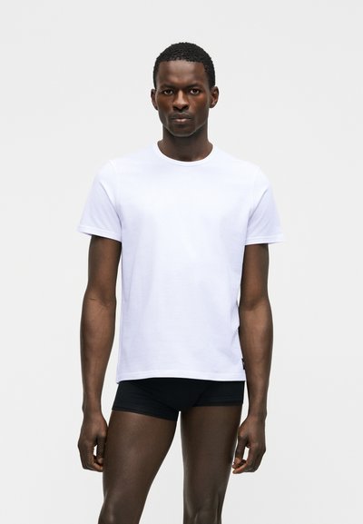 Νέος άνδρας που φοράει απλό λευκό κοντομάνικο μπλουζάκι και μαύρο σορτς, στέκεται μπροστά σε ανοιχτό γκρι φόντο, κοιτώντας μπροστά.