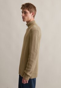 Strickpullover in hellem Beige mit hohem Kragen, strukturiertem Muster und gerippten Bündchen. Modell seitlich abgebildet, der Stoff wirkt weich.