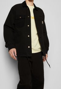 Svart, oversized jacka med knäppning och två bröstfickor; matchad med en ljusgul t-shirt. Tyg av texturerad bomull.