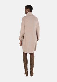 Abito in maglia beige oversize con colletto alto, spalle scese e maniche a palloncino, abbinato a stivali alti marroni con tacco.