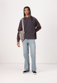 Brun og grå langærmet skjorte, lys blå flared jeans, sorte fritidssko, grå taske med lynlås, står på en hvid baggrund.