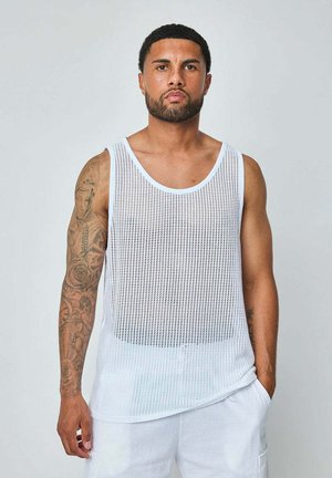 Homme aux cheveux courts et bouclés avec une barbe, portant un débardeur en maille blanche et un short blanc, debout avec une main dans la poche devant un fond uni.