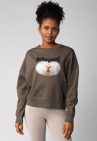 Vrouw met krullend haar die grote oorbellen draagt en een bruine sweatshirt aan heeft waarop een close-up van de neus van een wit dier staat afgebeeld, samen met het woord "Schnauze."