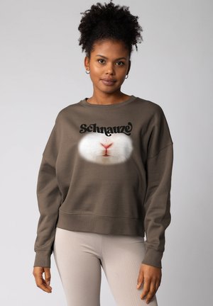 Femme avec des cheveux bouclés portant des boucles d'oreilles créoles et un sweat-shirt marron affichant un gros plan du nez d'un animal blanc et le mot "Schnauze."