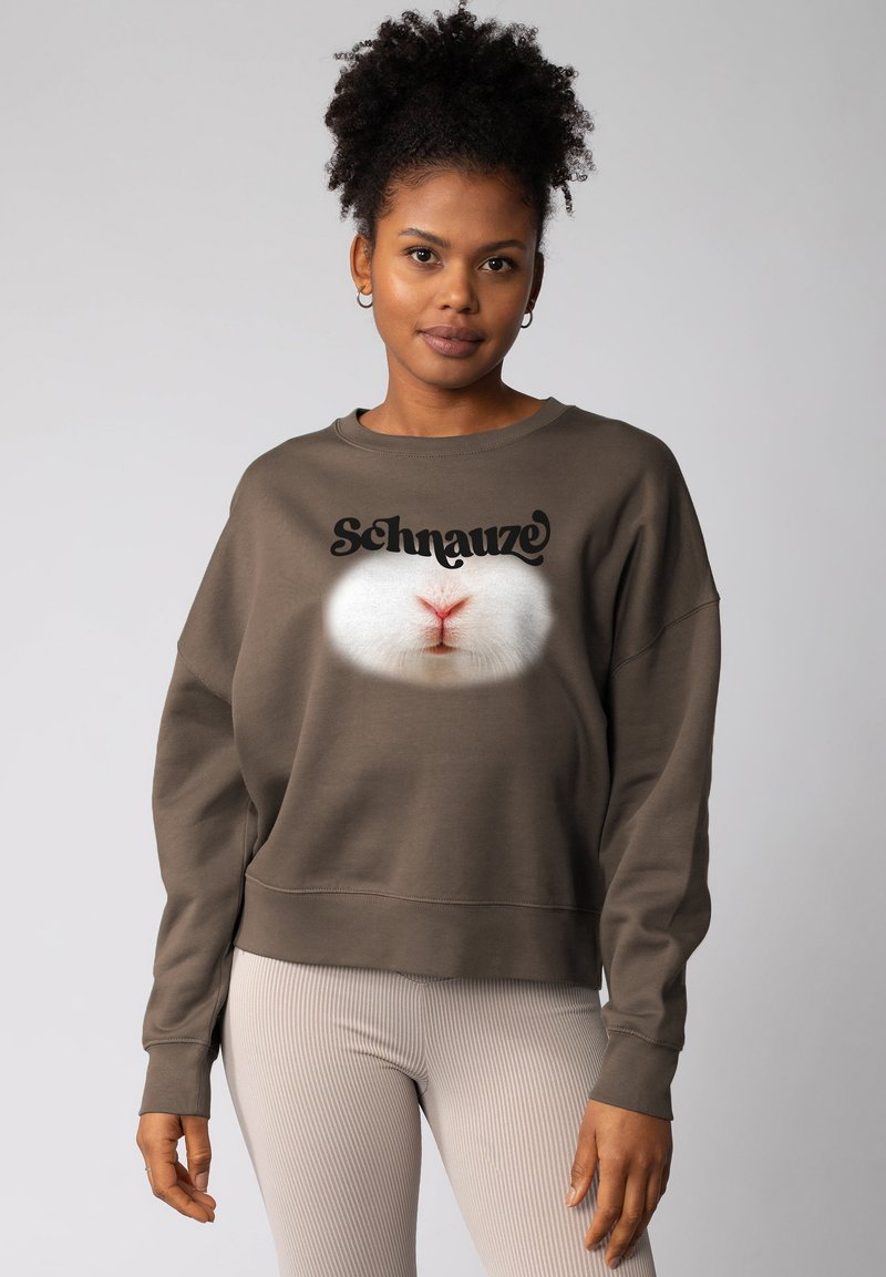 Vrouw met krullend haar die grote oorbellen draagt en een bruine sweatshirt aan heeft waarop een close-up van de neus van een wit dier staat afgebeeld, samen met het woord "Schnauze."