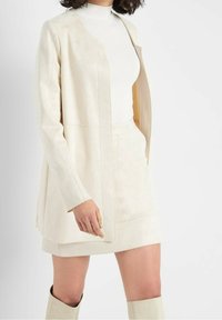 Veste couleur crème, aspect daim, au design ajusté, devant ouvert et poches latérales. Portée avec un col roulé blanc et une jupe ajustée.