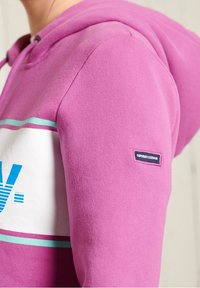 Sudadera con capucha morada con un diseño gráfico blanco y verde azulado, textura suave, con una etiqueta de logo en la manga y cordón ajustable.