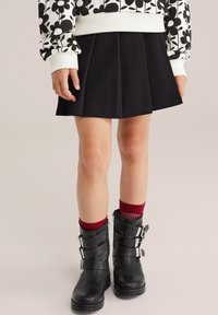 Kind in einem schwarzen Plisseerock, schwarzen Schnallenschuhen, roten Socken und einem weißen Sweatshirt mit schwarz floralem Muster, steht auf einem hellen Boden.