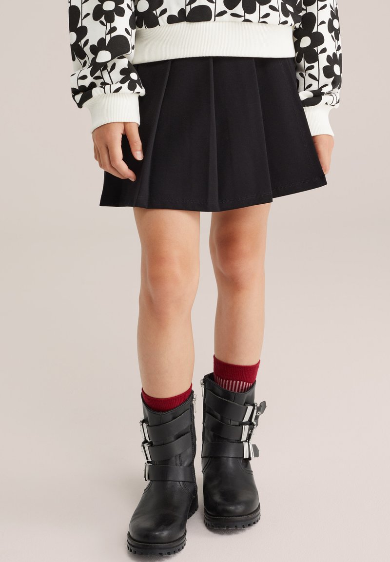 Kind in einem schwarzen Plisseerock, schwarzen Schnallenschuhen, roten Socken und einem weißen Sweatshirt mit schwarz floralem Muster, steht auf einem hellen Boden.