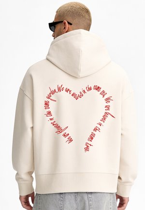 Person mit blondem Haar, die schwarze Sonnenbrille und einen weißen Hoodie mit rotem herzförmigem Text auf dem Rücken trägt.