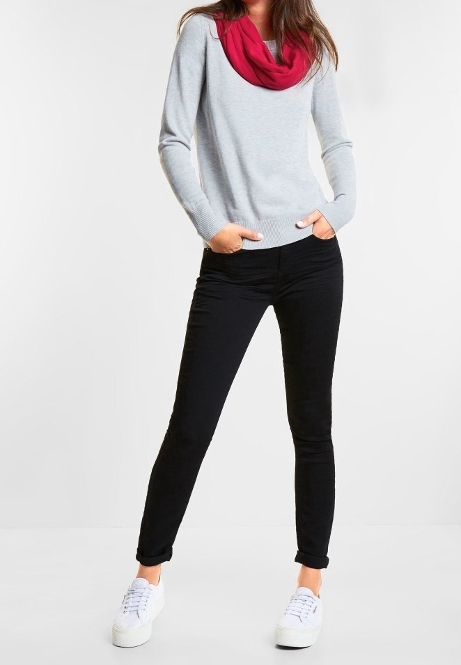 Femme debout les mains dans les poches, portant un pull gris clair, une écharpe rouge, un pantalon noir et des baskets blanches sur fond blanc.