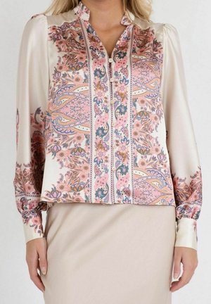 Vrouw die een blouse met lange mouwen draagt met roze en blauwe bloemige paisley patronen en een beige rok, getoond van schouders tot heupen.