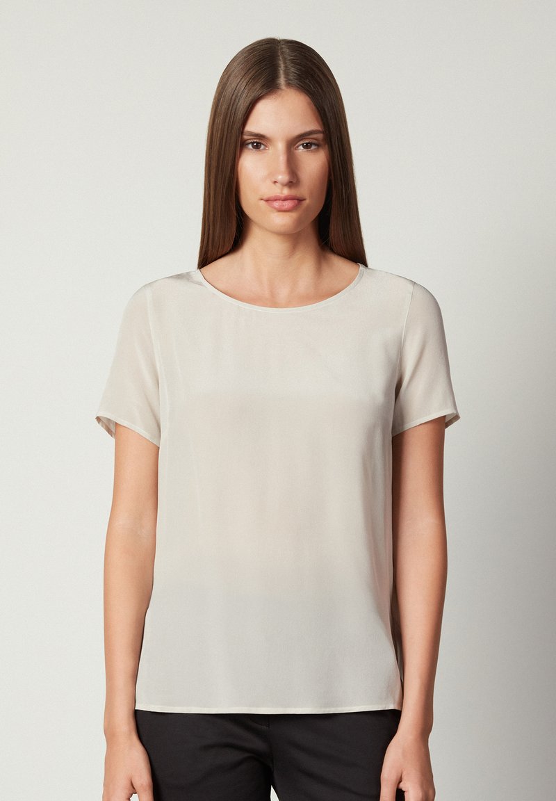 Kortärmad blus i ljusbeige, tillverkad av glatt tyg. Har en rund halsringning och en avslappnad passform. Inga synliga mönster eller metalldelar.