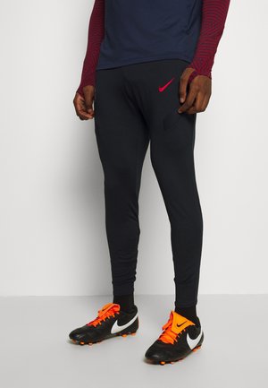 Muž v černých sportovních kalhotách s červeným logem Nike, v černých a oranžových kopačkách a tmavě modrém s červeným dlouhém rukávu, stojící na jednobarevném pozadí.