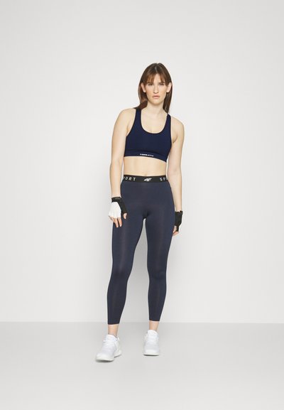 Navy sports bra with logo and high-waisted navy leggings; textured fabric; white athletic shoes; fingerless gloves.