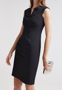 Svart ärmlös klänning med en figurnära silhuett, V-urringning och strukturerade sömmar. Accenterad med en silverclutch med ränder.