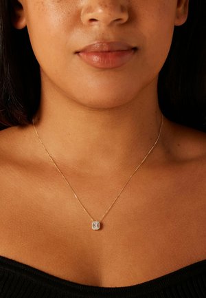 Gros plan sur le cou et la poitrine d'une femme, montrant un délicat collier en chaîne d'or avec un pendentif carré en diamant.