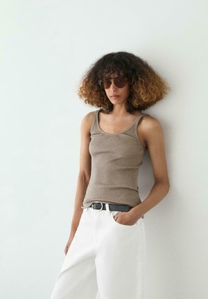 Vrouw met krullend ombre haar, zonnebril, een beige geribd tanktop, witte broek en een zwarte riem, leunend tegen een witte muur.