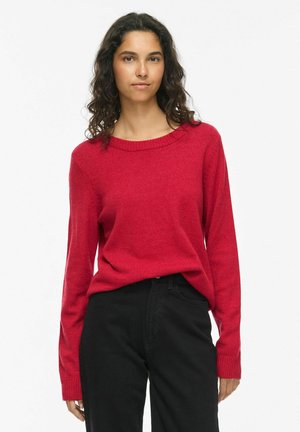 Femme aux longs cheveux bouclés et foncés, portant un pull rouge à manches longues et un pantalon noir, debout devant un fond blanc uni.