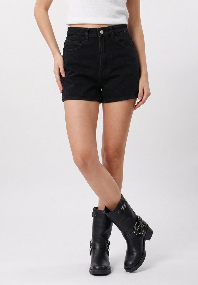 Kvinne iført svarte jeansshorts, svarte skinnstøvler med spenner som går til midt på leggen, og en hvit kort crop-top, stående mot en ensfarget bakgrunn.