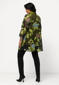 Zwarte doorzichtige kimono met een kleurrijk bloemenpatroon in groen, blauw en bordeauxrood. Heeft brede mouwen en een kraag, gedragen over zwarte broeken.
