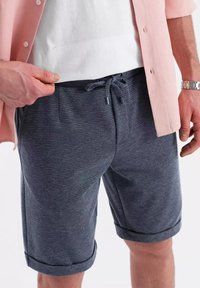 Shorts à motif marine avec une taille élastique, une fermeture à cordon et des ourlets repliés. Portés avec une chemise rose clair sur un tee-shirt blanc.