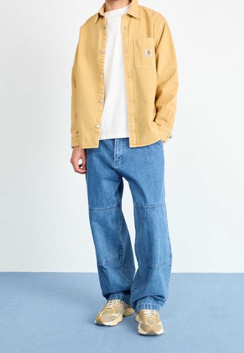Personne portant une chemise beige à boutons, un t-shirt blanc, un jean bleu ample et des baskets beiges debout sur un sol bleu avec un fond blanc.