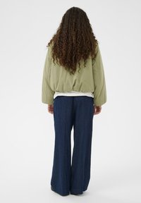 Zeleno podloženo jopico z zaobljenim robom, nošeno čez belo srajco, v kombinaciji z širokimi temnimi denim hlačami, ki imajo gladko teksturo.